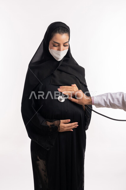 الالتزام بالاحترازات الوقائيه ضد كوفيد 19، الاهتمام بصحة الجنين، طبيبة عربية خليجية سعودية ترتدي المعطف الطبي تضع السماعات الطبية على بطن إمرأة حامل، بورتريه مقرب لشابة إماراتية ترتدي القناع الوقائي تنظر إلى الكاميرا، تقديم الاستشارات الطبية، خلفية بيضاء