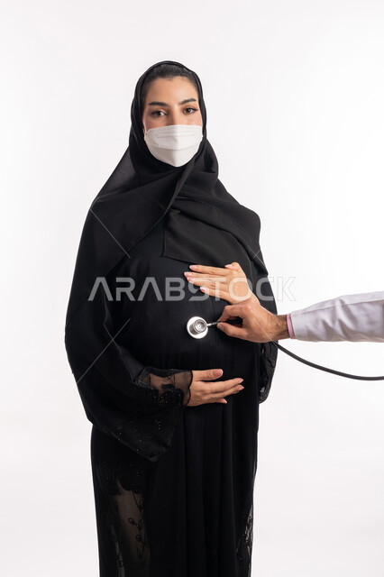 الاهتمام بصحة الجنين، الالتزام بالاحترازات الوقائيه ضد كوفيد 19، طبيبة عربية خليجية سعودية ترتدي المعطف الطبي تضع السماعات الطبية على بطن إمرأة حامل، بورتريه مقرب لشابة إماراتية ترتدي القناع الوقائي تنظر إلى الكاميرا، تقديم الاستشارات الطبية، خلفية بيضاء