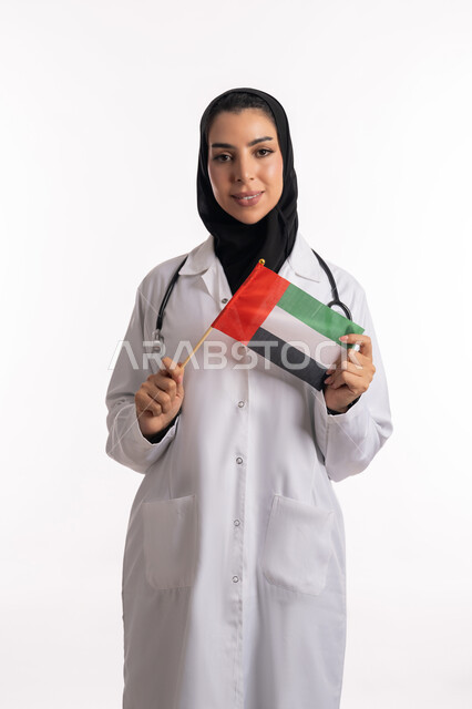 العمل في المستشفيات والمراكز الطبية، بورتريه مقرب لشابة إماراتية تحتفل باليوم الوطني لدولة الامارات العربية المتحدة تحمل بيدها علم دولة الامارات، طبيبة عربية خليجية سعودية محجبة ترتدي معطف الطبيب تنظر إلى الكاميرا بإيماءات الحب والسعادة، خلفية بيضاء