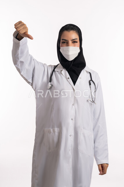 الإنزعاج من شيء ما، الالتزام بالاحترازات الوقائيه ضد كوفيد 19، طبيبة عربية خليجية سعودية ترتدي المعطف الطبي وسماعة الأذن تشير بإصبع الإبهام للأسفل بإيماءات عدم الموافقة والرفض، بورتريه مقرب لشابة إماراتية ترتدي الكمامة تنظر إلى الكاميرا، خلفية بيضاء