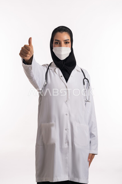 ارتداء الكمامة للوقاية من العدوى، الالتزام بالاحترازات الوقائيه ضد كوفيد 19، طبيبة عربية خليجية سعودية ترتدي المعطف الطبي وسماعة الأذن تشير بإصبع الإبهام للأعلى، بورتريه مقرب لشابة إماراتية تنظر إلى الكاميرا، الرضا والقبول بجوده الخدمات الطبية، خلفية بيضاء