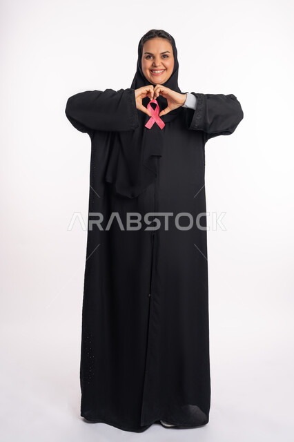 الكشف المبكر عن سرطان الثدي، أثر الدعم الايجابي على الصحة الجسدية، بورتريه لإمرأة عربية خليجية إماراتيه ترتدي الحجاب والعباءة تضع الشريط الوردي تقوم بتشكيل اليدين على شكل قلب، سيدة سعودية تنظر إلى الكاميرا بتعابير الحب والتعاطف، صورة لكامل الجسم، خلفية بيضاء