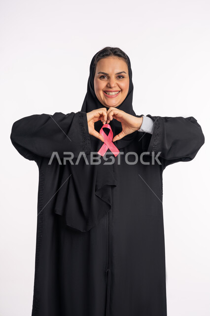 أثر الدعم الايجابي على الصحة النفسية والجسدية، بورتريه لإمرأة عربية خليجية إماراتيه ترتدي الحجاب والعباءة تضع الشريط الوردي تقوم بتشكيل اليدين على شكل قلب، سيدة سعودية تنظر إلى الكاميرا بتعابير الحب والتعاطف، أهمية الوعية بالكشف المبكر عن سرطان الثدي، خلفية بيضاء