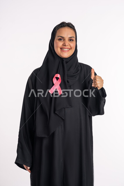 حملات الإرشاد والتنبيه بالكشف المبكر عن سرطان الثدي، شابة سعودية محجبة مبتسمة تقوم برفع يدها بإيماءات الإعجاب والرضا، بورتريه لامرأة عربية خليجية إماراتيه ترتدي العباية تضع على صدرها شريط وردي للتوعية بالأمراض الخبيثة، الحفاظ على الصحة الجسدية، خلفية بيضاء