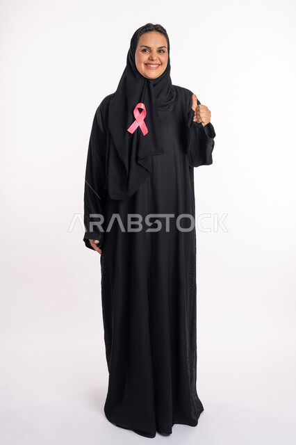 سيدة سعودية محجبة مبتسمة تقوم برفع يدها بإيماءات الإعجاب والرضا، حملات الإرشاد والتنبيه بالكشف المبكر عن سرطان الثدي، بورتريه لامرأة عربية خليجية إماراتيه ترتدي العباية تضع على صدرها شريط وردي للتوعية بالأمراض الخبيثة، صورة لكامل الجسم، خلفية بيضاء