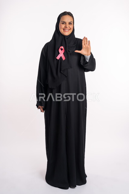 طلب التوقف عن إهمال الصحة الجسدية، شابة سعودية تضع شعار الحملة التوعوية بضرورة الكشف المبكر عن سرطان الثدي، بورتريه لإمرأة عربية خليجية إماراتية مبتسمة ترتدي الحجاب والعباءة والشريط الوردي ترفع يدها بحركات الرفض والقوة، صورة الجسم بالكامل، خلفية بيضاء