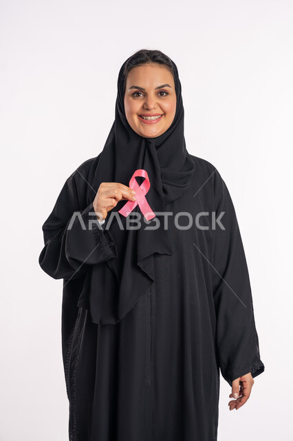 تحدي الأمراض الخبيثة، رمز الحملة العالمية، بورتريه لإمرأة عربية خليجية إماراتية ترتدي الحجاب والعباءة السوداء تحمل بيديها الشريط الوردي، شابة سعودية تنظر الى الكاميرا بإيماءات البهجة والتفاؤل، حملة نسائية توعوية للكشف المبكر عن سرطان الثدي، خلفية بيضاء
