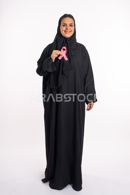حملة نسائية توعوية للكشف المبكر عن سرطان الثدي، تحدي الأمراض الخبيثة، بورتريه لإمرأة عربية خليجية إماراتية ترتدي الحجاب والعباءة السوداء تحمل بيديها الشريط الوردي، شابة سعودية تنظر الى الكاميرا بإيماءات البهجة والتفاؤل، صورة لكامل الجسم، خلفية بيضاء
