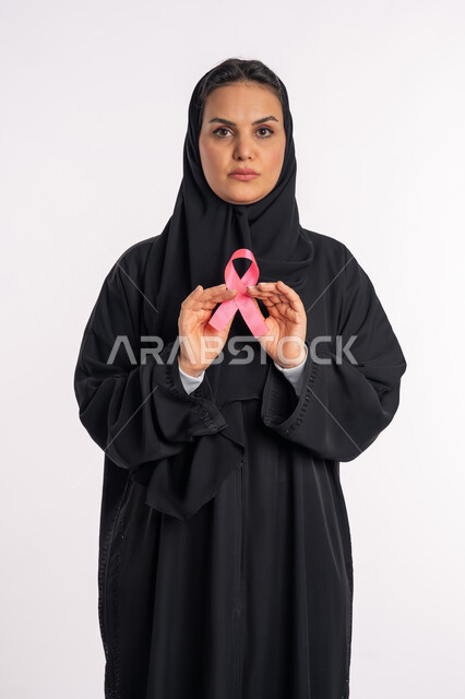 التوعية بضرورة الكشف المبكر عن سرطان الثدي، شابة سعودية تنظر الى الكاميرا بإيماءات القوة والصمود، تحدي الأمراض الخبيثة، بورتريه لإمرأة عربية خليجية إماراتية ترتدي الحجاب والعباءة السوداء تحمل بيديها الشريط الوردي، خلفية بيضاء
