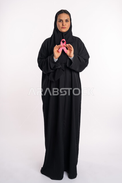 A young Saudi woman looks at the camera with gestures of strength and defiance, a women’s awareness campaign for early detection of breast cancer, the challenge of malignant diseases, a portrait of an Arab Gulf Emirati woman wearing the hijab and black abaya, holding a pink ribbon in her hands, full-body portrait, white background