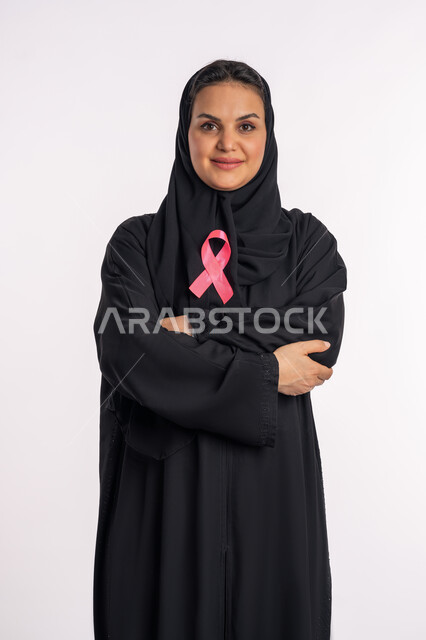 شابة سعودية تقف باستقامة وتنظر إلى الكاميرا بإيماءات البهجة والتفاؤل، شعار الحملة العالمية، اليوم العالمي للكشف المبكر عن سرطان الثدي، بورتريه لإمرأة عربية خليجية إماراتية ترتدي الحجاب والعباءة السوداء تقوم باحتضان الشريط الزهري، خلفية بيضاء