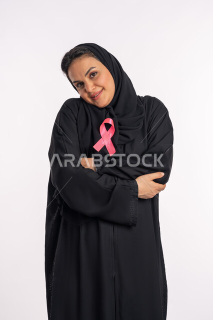 تحدي الأمراض الخبيثة، شعار الحملة العالمية، اليوم العالمي للكشف المبكر عن سرطان الثدي، بورتريه لإمرأة عربية خليجية إماراتية ترتدي الحجاب والعباءة السوداء تقوم باحتضان الشريط الزهري، شابة سعودية تميل رأسها بإيماءات البهجة والتفاؤل، خلفية بيضاء