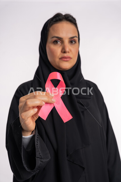 World Breast Cancer Awareness Day, looking at the camera with gestures of sadness and depression, early detection of the disease and attention to its treatment, a Saudi woman holding the symbol of the global campaign, a portrait of an Arab Gulf Emirati woman wearing a hijab and abaya and holding a pink ribbon in her hand, a pink background