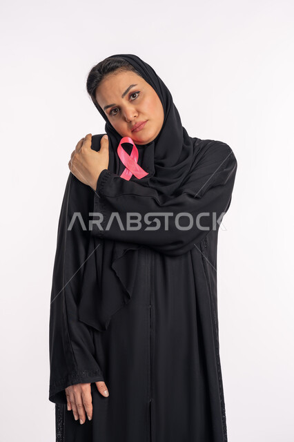 شابة سعودية تضع يدها على كتفها وتميل رأسها بإيماءات الحزن والاكتئاب، الإصابة بالمرض الخبيث، بورتريه لإمرأة عربية خليجية إماراتية ترتدي الحجاب والعباءة السوداء تقوم باحتضان شعار الكشف المبكر عن سرطان الثدي، خلفية بيضاء