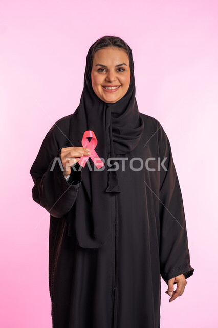 الكشف المبكر عن المرض والاهتمام بعلاجه ،شابة سعودية تمسك برمز الحملة العالمية، بورتريه لامرأة عربية خليجية إماراتية مبتسمة ترتدي الحجاب والعباءة وتحمل بيدها شريط زهري اللون، اليوم العالمي للتوعية بسرطان الثدي، النظر إلى الكاميرا بإيماءات السرور، خلفية وردية
