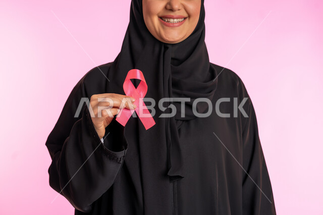 اليوم العالمي للتوعية بسرطان الثدي، الكشف المبكر عن المرض والاهتمام بعلاجه ،شابة سعودية تمسك برمز الحملة العالمية، بورتريه لامرأة عربية خليجية إماراتية مبتسمة ترتدي الحجاب والعباءة وتحمل بيدها شريط زهري اللون، خلفية وردية