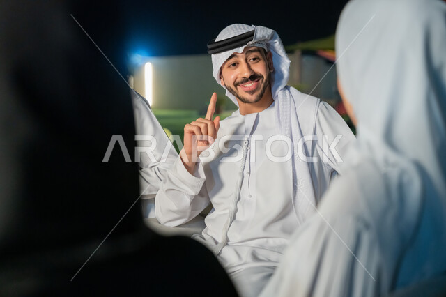 ايماءات البهجة والسرور، أجواء ليلية هادئة، شاب عربي خليجي إماراتي يرتدي الكندورة والغترة يقوم بتبادل الأحاديث الشيقة مع الأصدقاء، الديكورات الخارجية الراقية، قضاء أوقات ممتعة مع الأقرباء، جلسات مسائية في شرفة المنزل