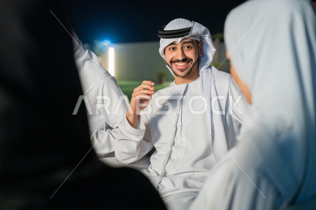 أجواء ليلية هادئة، شاب عربي خليجي إماراتي يرتدي الكندورة والغترة يقوم بتبادل الأحاديث الشيقة مع الأصدقاء، ايماءات البهجة والسرور، الديكورات الخارجية الراقية، قضاء أوقات ممتعة مع الأقرباء، جلسات مسائية في شرفة المنزل