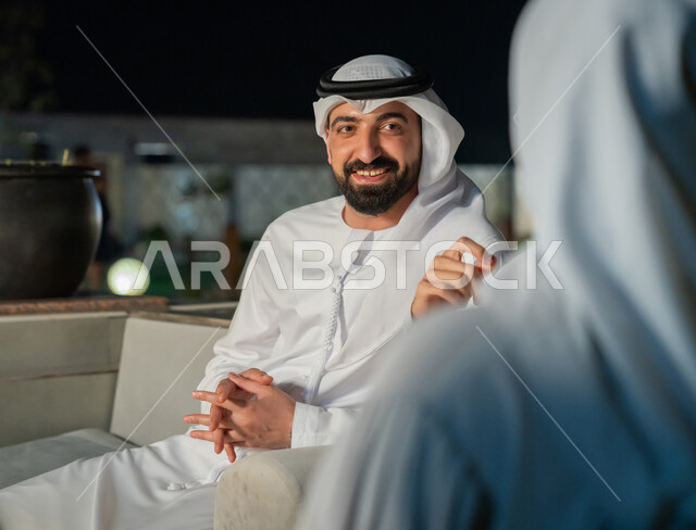 جلسات مسائية في شرفة المنزل، ايماءات البهجة والسرور، أجواء ليلية هادئة، شاب عربي خليجي إماراتي يرتدي الكندورة والغترة يقوم بتبادل الأحاديث الشيقة، الديكورات الخارجية الراقية، قضاء أوقات ممتعة مع الأصدقاء،