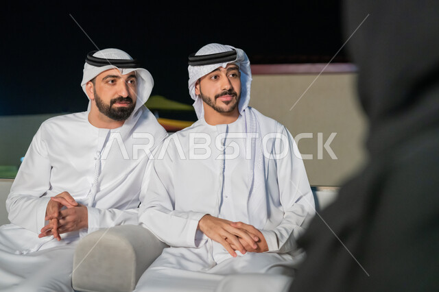 جلسات مسائية في شرفة المنزل، شابان عربيان خليجيان إماراتيان يرتديان الكندورة والغترة يتبادلان الأحاديث الشيقة مع الأصدقاء، أجواء ليلية هادئة، بايماءات البهجة والسرور، الديكورات الخارجية الراقية، قضاء أوقات ممتعة مع الأقرباء