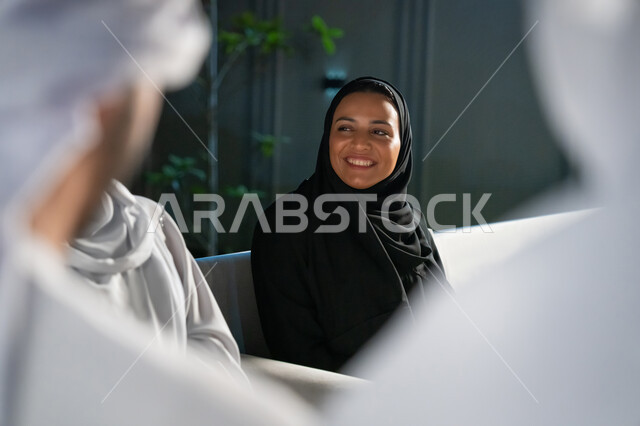 تبادل الأحاديث الشيقة بايماءات البهجة والسرور، الديكورات الخارجية الراقية، أجواء ليلية هادئة، صورة مقربة لشابة عربية خليجية إماراتية محجبة ترتدي العباية تجلس مع مجموعة من الأصدقاء في شرفة منزل، قضاء أوقات ممتعة مع الأقرباء