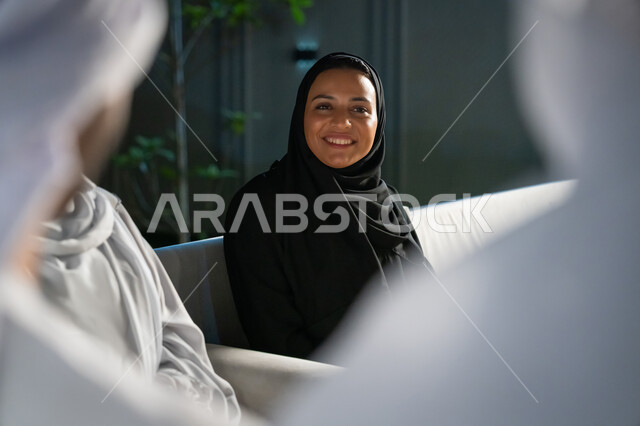 ايماءات البهجة والسرور، الديكورات الخارجية الراقية، تبادل الأحاديث الشيقة، أجواء ليلية هادئة، صورة مقربة لشابة عربية خليجية إماراتية محجبة ترتدي العباية تجلس مع مجموعة من الأصدقاء في شرفة منزل، قضاء أوقات ممتعة مع الأقرباء