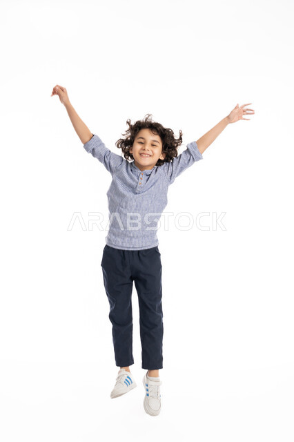 The concept of encouragement and enthusiasm, a portrait of an Arab Gulf Emirati boy wearing a casual outfit, jumping enthusiastically and raising his hands up, joy in victory and triumph, a smiling Saudi boy with expressions and gestures of success and achieving goals, full-length body portrait, white background.