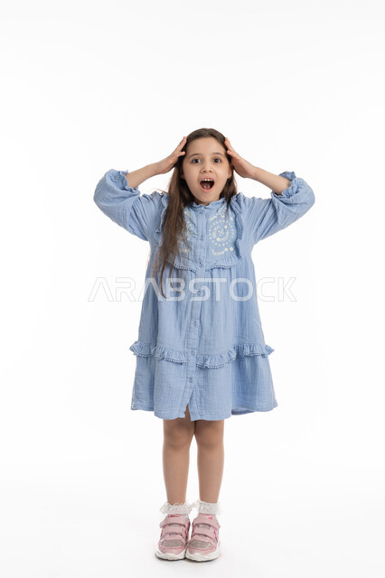 الذهول و الصدمة والتفاجؤ من شيئ ما, بورتريه لفتاة عربية امارتية خليجية بتعابير وجه ويدين تدل على الدهشه و التعجب, بنت سعودية ترتدي الفستان وترفع يديها على رأسها بإيماءات الفرح والسعادة، صورة الجسم بالكامل، خلفية بيضاء