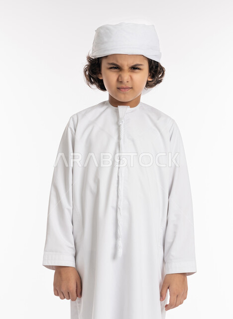 الغضب من شيء ما, صبي سعودي يقف بايماءات الضجر و الاستياء وعدم الرضا, بورتريه مقرب لولد عربي اماراتي خليجي يرتدي الكندورة والعمامة ينظر الى الكاميرا بعصبية, خلفية بيضاء
