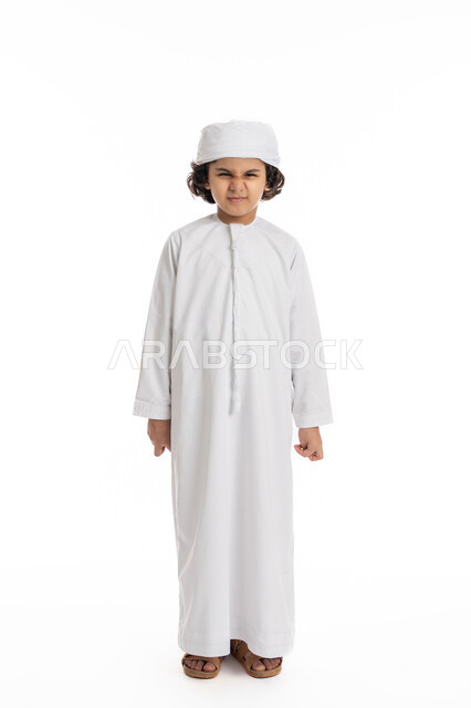 الضجر و الاستياء وعدم الرضا، صبي سعودي يقف بايماءات الغضب من شيء ما, بورتريه لولد عربي اماراتي خليجي يرتدي الكندورة والعمامة ينظر الى الكاميرا بعصبية, صورة الجسم بالطول الكامل، خلفية بيضاء