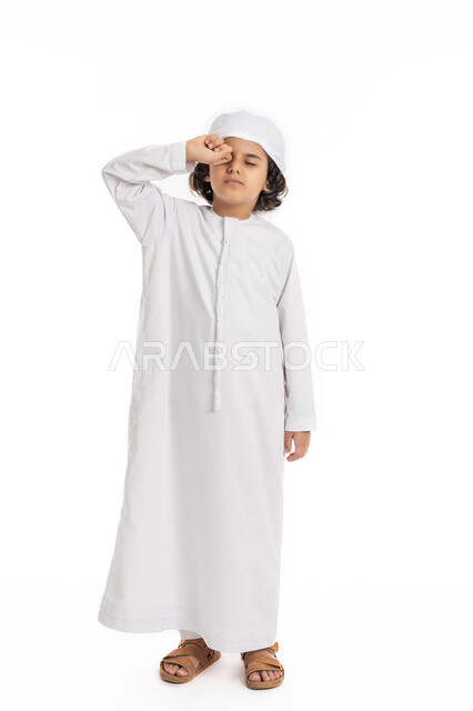 الشعور بالتعب والكسل والخمول, صبي سعودي يقوم بفرك عينه، الإحساس بالإرهاق و الحاجه الى النوم والراحه ,بورتريه لولد عربي اماراتي خليجي يرتدي الكندورة والعمامة يقف بإيماءات تدل على النعاس, صورة الجسم بالكامل، خلفية بيضاء
