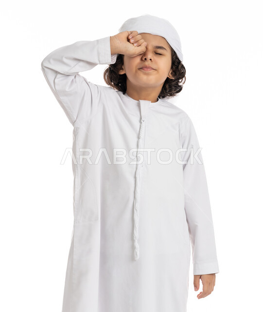 الإحساس بالإرهاق و الحاجه الى النوم والراحه ,بورتريه مقرب لولد عربي اماراتي خليجي يرتدي الكندورة والعمامة يقف بإيماءات تدل على النعاس, الشعور بالتعب والكسل والخمول, صبي سعودي يقوم بفرك عينه، خلفية بيضاء