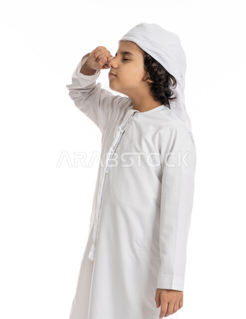 الإحساس بالإرهاق و الحاجه الى النوم والراحه ,بورتريه مقرب لولد عربي اماراتي خليجي يرتدي الكندورة والعمامة يقف بإيماءات تدل على النعاس, الشعور بالتعب والكسل والخمول, صبي سعودي يقوم بفرك عينه، خلفية بيضاء
