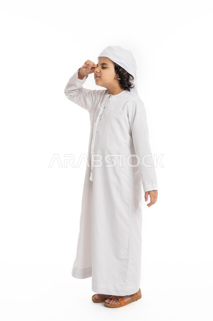 الشعور بالتعب والكسل والخمول, صبي سعودي يقوم بفرك عينه، الإحساس بالإرهاق و الحاجه الى النوم والراحه ,بورتريه لولد عربي اماراتي خليجي يرتدي الكندورة والعمامة يقف بإيماءات تدل على النعاس, صورة الجسم بالكامل، خلفية بيضاء