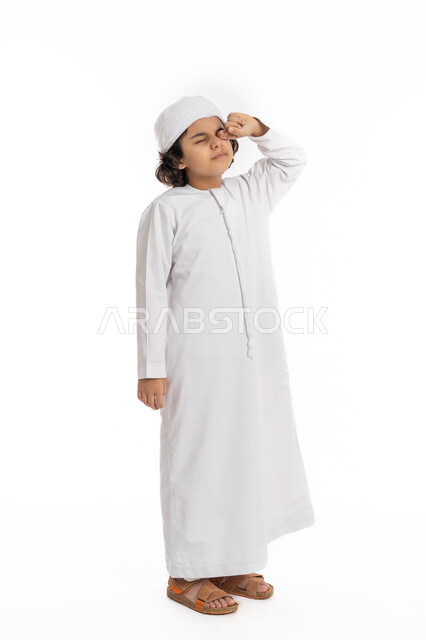 Feeling exhausted and needing sleep and rest, portrait of an Arab Emirati Gulf boy wearing a kandora and turban, standing with gestures indicating sleepiness, feeling tired, lazy and lethargic, a Saudi boy rubbing his eyes, full body portrait, white background