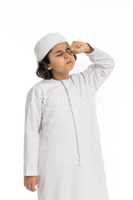 الإحساس بالإرهاق و الحاجه الى النوم والراحه ,بورتريه مقرب لولد عربي اماراتي خليجي يرتدي الكندورة والعمامة يقف بإيماءات تدل على النعاس, الشعور بالتعب والكسل والخمول, صبي سعودي يقوم بفرك عينه، خلفية بيضاء