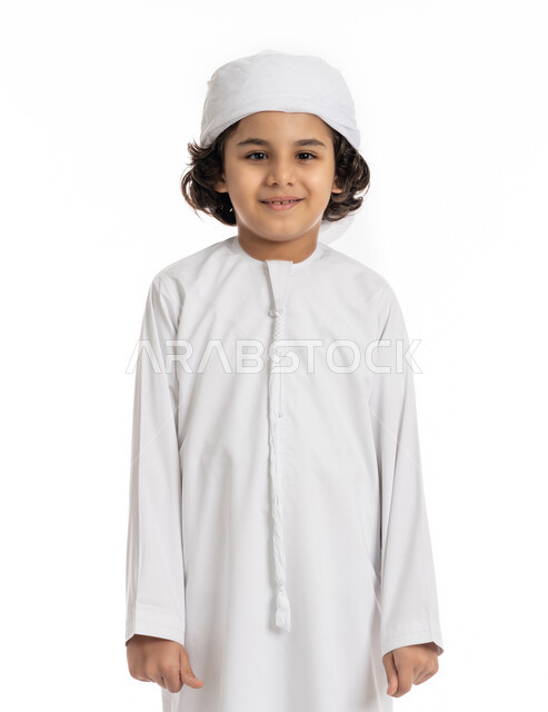 فتى سعودي يقف باستقامة بتعابير وايماءات الجدية والثقة بالنفس، بورتريه مقرب لولد عربي اماراتي خليجي مبتسم يرتدي االعمامة والكندورة ينظر الى الكاميرا بإيماءات الحزم والسرور، الاهتمام بالأناقة والمظهر الخارجي، خلفية بيضاء