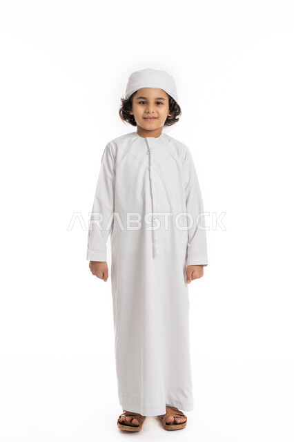 الاهتمام بالأناقة والمظهر الخارجي، فتى سعودي يقف باستقامة بتعابير وايماءات الجدية والثقة بالنفس، بورتريه لولد عربي اماراتي خليجي مبتسم يرتدي االعمامة والكندورة ينظر الى الكاميرا بإيماءات الحزم والسرور، صورة الجسم بالكامل، خلفية بيضاء