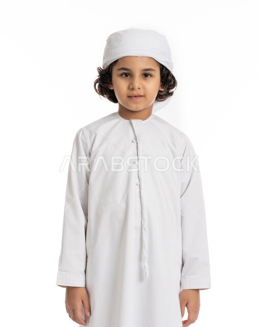 فتى سعودي يقف باستقامة بتعابير وايماءات الجدية والثقة بالنفس، بورتريه مقرب لولد عربي اماراتي خليجي مبتسم يرتدي االعمامة والكندورة ينظر الى الكاميرا بإيماءات الحزم والسرور، الاهتمام بالأناقة والمظهر الخارجي، خلفية بيضاء