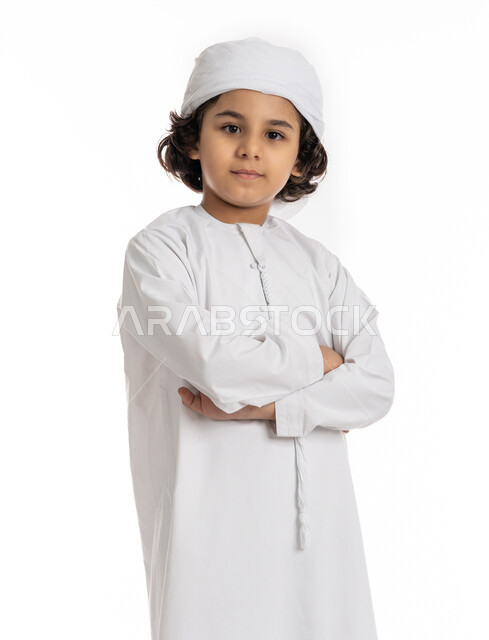 صبي سعودي يقف بأيدي متقاطعة بتعابير الثقة بالنفس، بورتريه مقرب لولد عربي خليجي إماراتي مبتسم يرتدي العمامة والكندورة ينظر إلى الكاميرا يإيماءات السرور، التقاط صور شخصية للوثائق الرسمية، الأناقه والاهتمام بالمظهر الخارجي، خلفية بيضاء