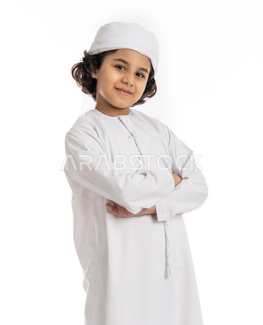 صبي سعودي يقف بأيدي متقاطعة بتعابير الثقة بالنفس، بورتريه مقرب لولد عربي خليجي إماراتي مبتسم يرتدي العمامة والكندورة ينظر إلى الكاميرا يإيماءات السرور، التقاط صور شخصية للوثائق الرسمية، الأناقه والاهتمام بالمظهر الخارجي، خلفية بيضاء