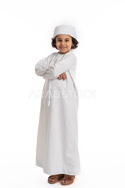 التقاط صور شخصية للوثائق الرسمية، الأناقه والاهتمام بالمظهر الخارجي، صبي سعودي يقف بأيدي متقاطعة بتعابير الثقة بالنفس، بورتريه لولد عربي خليجي إماراتي مبتسم يرتدي العمامة والكندورة ينظر إلى الكاميرا يإيماءات السرور، صورة الجسم بالكامل، خلفية بيضاء