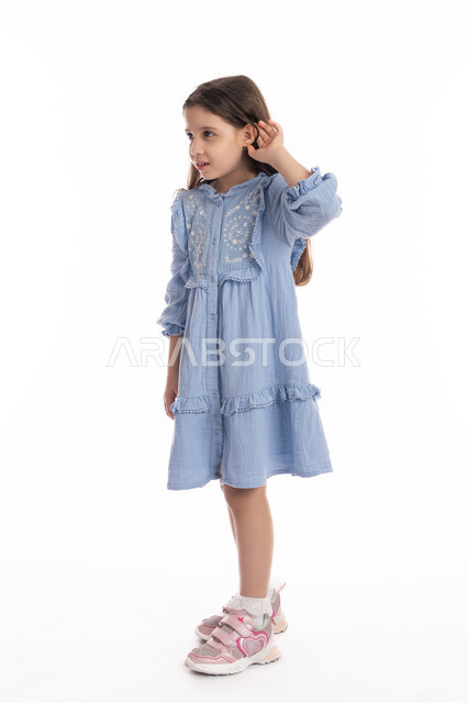 Hearing conversations and discussions, trying to spy and eavesdrop, gestures of listening and revealing secrets, a portrait of an Arab Emirati Gulf girl wearing a dress, putting her hands behind her ears, a Saudi girl listening and focusing on the words, full body image, white background