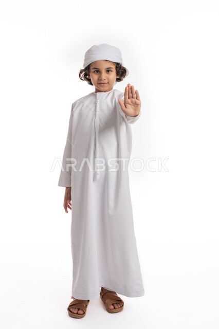 الصمت التوقف عن الكلام، الشعور بالإنزعاج و الرفض والإمتناع، فتى سعودي يقف بإيماءات يدين تدل على الأمر بالتوقف عن فعل شيء ما و النهي عنه، بورتريه لولد عربي اماراتي خليجي يرتدي الكندورة والعمامة الاماراتية ويرفع كفه للأعلى, صورة الجسم بالكامل، خلفية بيضاء
