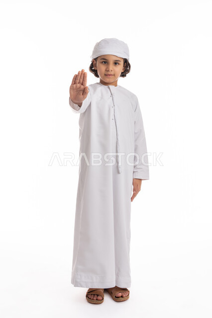 الصمت التوقف عن الكلام، الشعور بالإنزعاج و الرفض والإمتناع، فتى سعودي يقف بإيماءات يدين تدل على الأمر بالتوقف عن فعل شيء ما و النهي عنه، بورتريه لولد عربي اماراتي خليجي يرتدي الكندورة والعمامة الاماراتية ويرفع كفه للأعلى, صورة الجسم بالكامل، خلفية بيضاء
