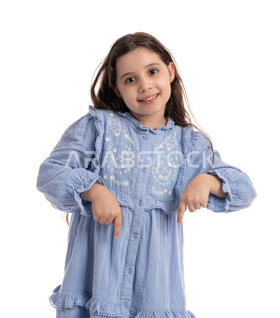 Two hand gestures indicating a specific location, specifying this place (here), a close-up portrait of an Arab Emirati Gulf girl pointing with her fingers downward, a smiling Saudi girl wearing a dress documenting this moment (now), a white background.