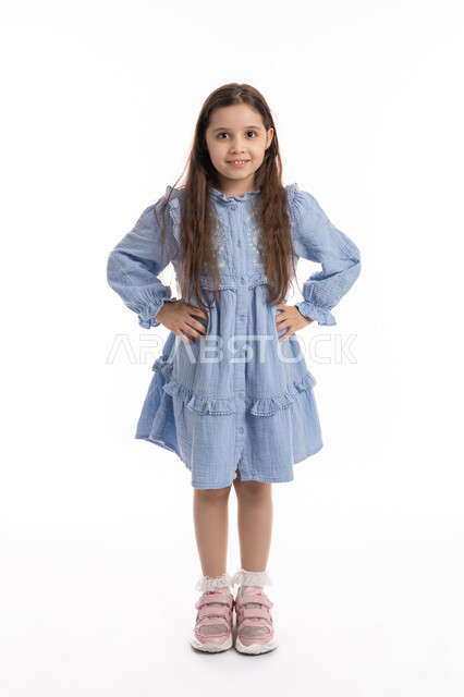 مفهوم الأنوثة والنعومة، بورتريه مقرب لفتاة عربية خليجية إماراتية مبتسمة ترتدي الفستان تنظر الى الكاميرا بفرح تضع يديها على خصرها، بنت سعودية تقف بتعابير وايماءات السعادة والسرور، خلفية بيضاء