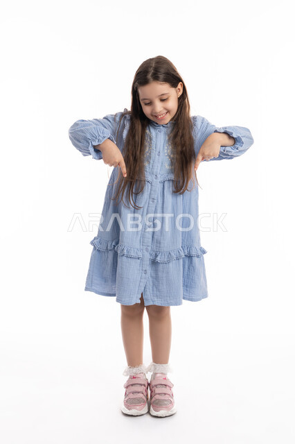 إيماءات يدين تشير الى موقع معين، تحديد هذا المكان (هنا), بورتريه لفتاة عربية اماراتية خليجية تشير بأصابعها إلى الاسفل, بنت سعودية مبتسمة ترتدي الفستان تقوم في توثيق هذه اللحظه (الآن)، صورة الجسم بالكامل، خلفية بيضاء