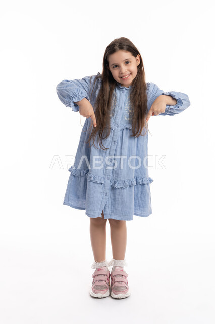 إيماءات يدين تشير الى موقع معين، تحديد هذا المكان (هنا), بورتريه لفتاة عربية اماراتية خليجية تشير بأصابعها إلى الاسفل, بنت سعودية مبتسمة ترتدي الفستان تقوم في توثيق هذه اللحظه (الآن)، صورة الجسم بالكامل، خلفية بيضاء