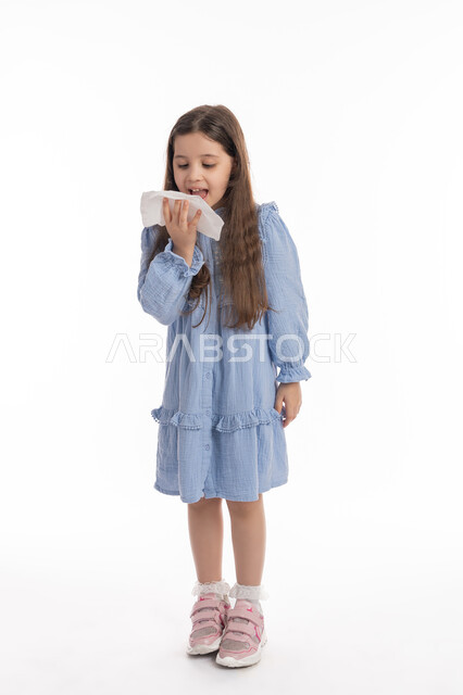 Using tissues to sneeze, feeling sick and cold, the need for treatment and medicine, a Saudi girl wearing a dress feeling tired and exhausted, a portrait of an Arab Emirati Gulf girl with facial and hand gestures indicating sneezing, full body portrait, white background
